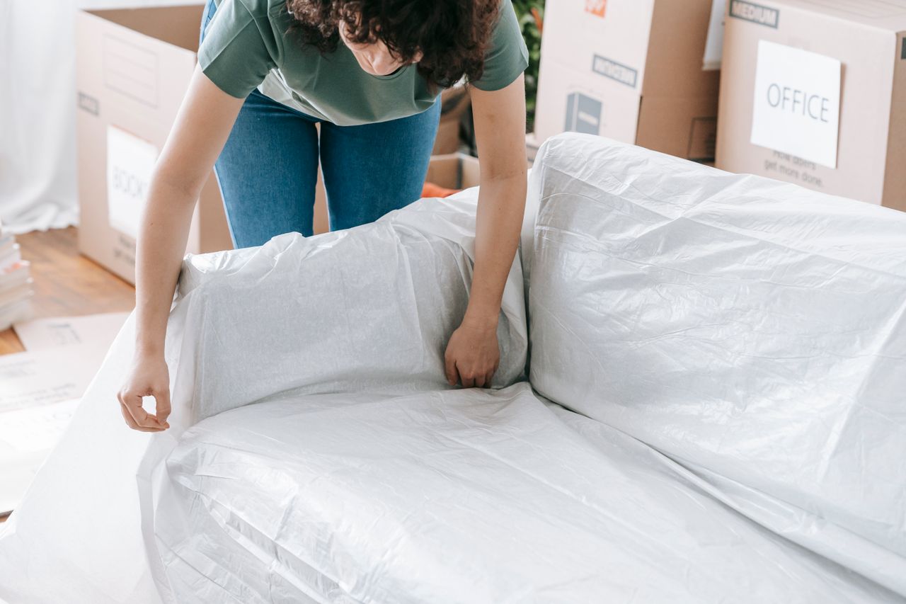Une femme enveloppe minutieusement un canapé avec une matière protectrice tout en préparant des cartons dans une pièce lumineuse.
